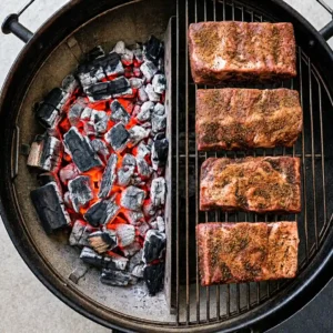 Top-down view of four seasoned beef back rib racks cooking on a round charcoal grill, set up for two-zone indirect grilling with glowing coals on the left side.