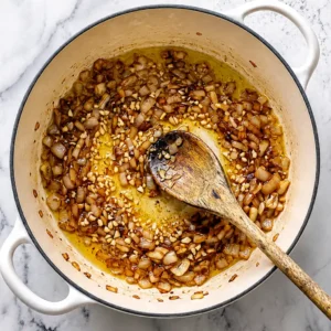 Top-down view of finely chopped onions and minced garlic sautéing until golden brown in oil inside a white Dutch oven, stirred with a wooden spoon.