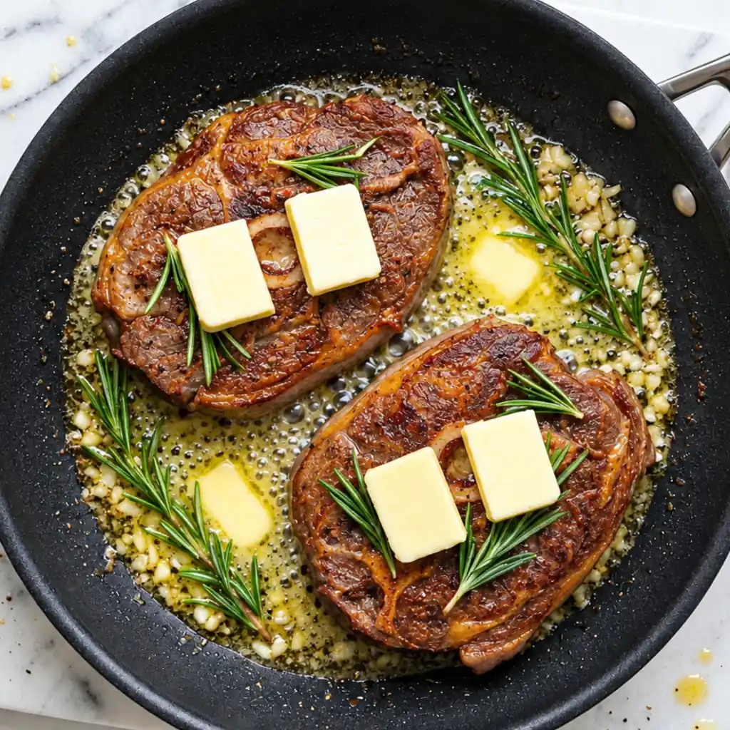Two bone-in lamb shoulder chops cooking in a skillet, topped with pat butter slices and fresh rosemary sprigs, surrounded garlic butter.