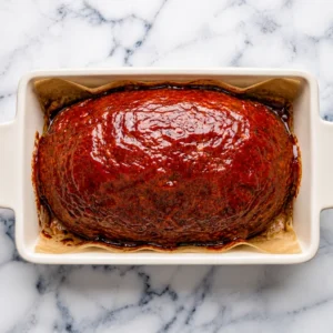 meatloaf seasoned with onion soup mix, coated in a glossy, rich tomato glaze, sitting in a parchment-lined white baking dish on a marble surface.