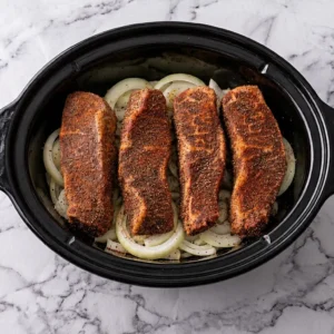 Four heavily seasoned country-style beef ribs arranged neatly on a bed of sliced onions inside a black slow cooker insert, set on a white marble countertop.