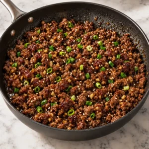 Savoury ground beef bulgogi simmering in a black skillet, garnished with chopped green onions and toasted sesame seeds on a marble countertop.