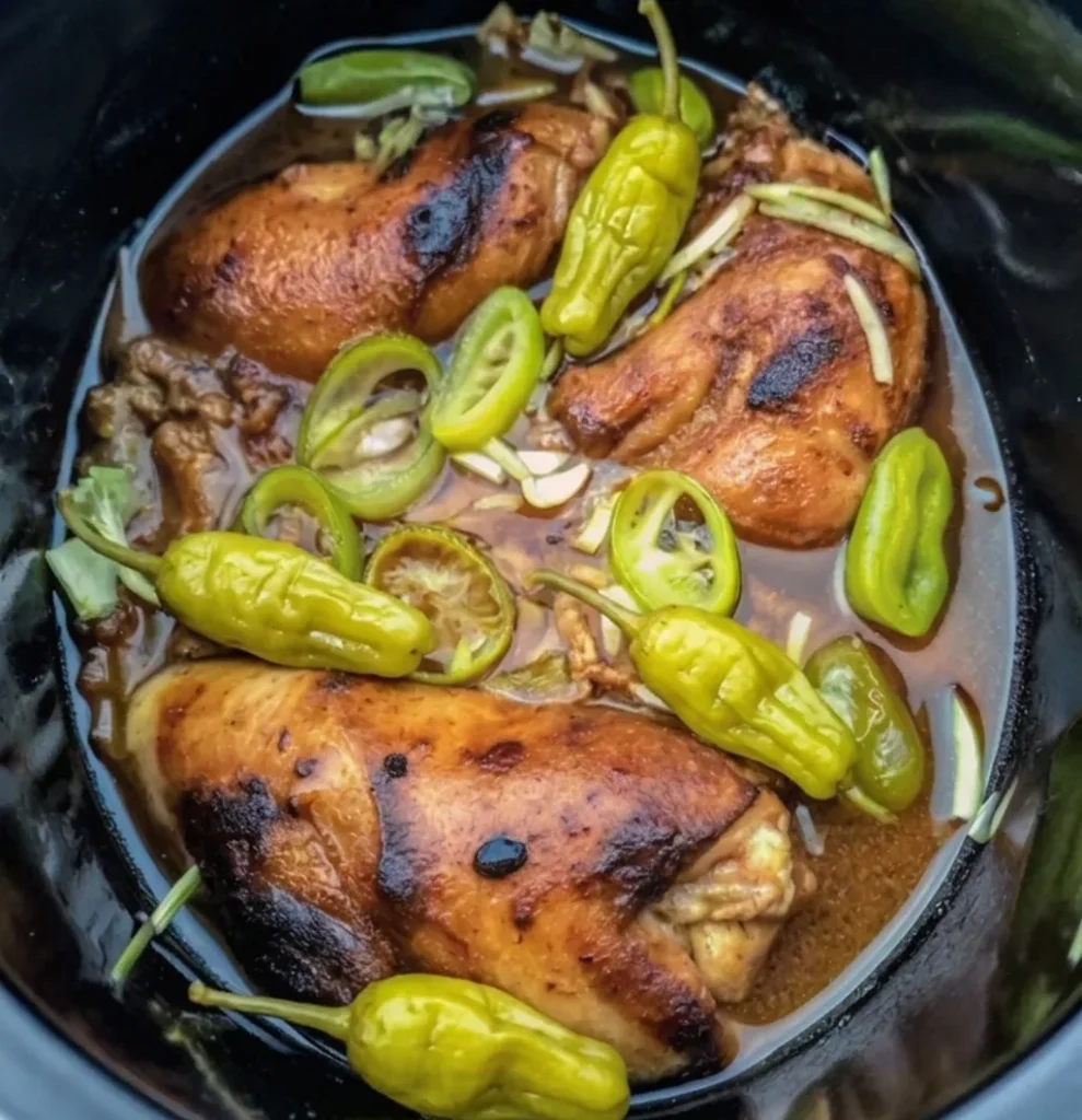 Cooked, browned Mississippi chicken breasts simmering in a rich gravy inside a slow cooker, topped with whole and sliced green pepperoncini peppers.
