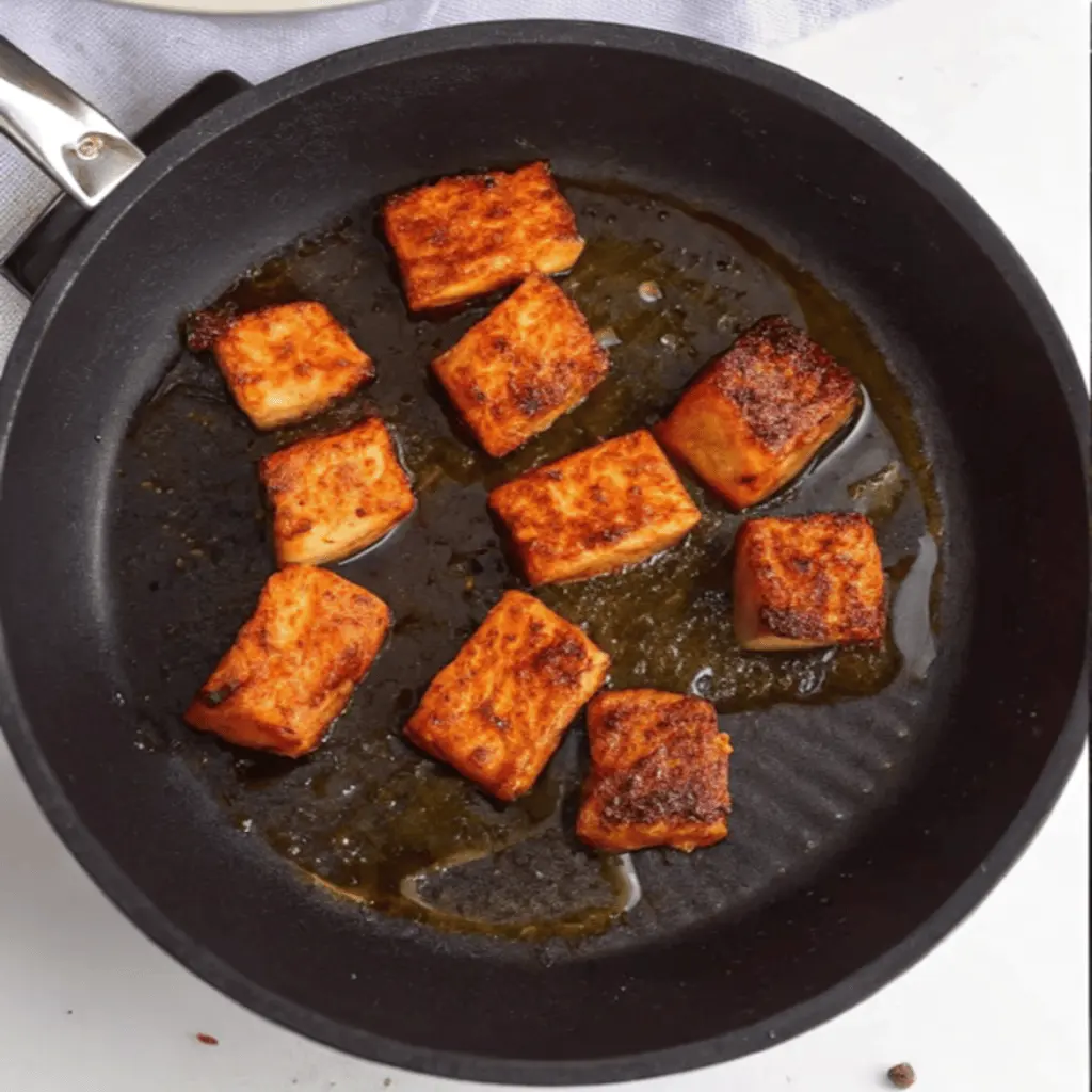 
A high-angle shot shows several seasoned salmon cubes frying in a black skillet with a small amount of oil. The salmon pieces are golden-brown and have a crispy, seared exterior from cooking.