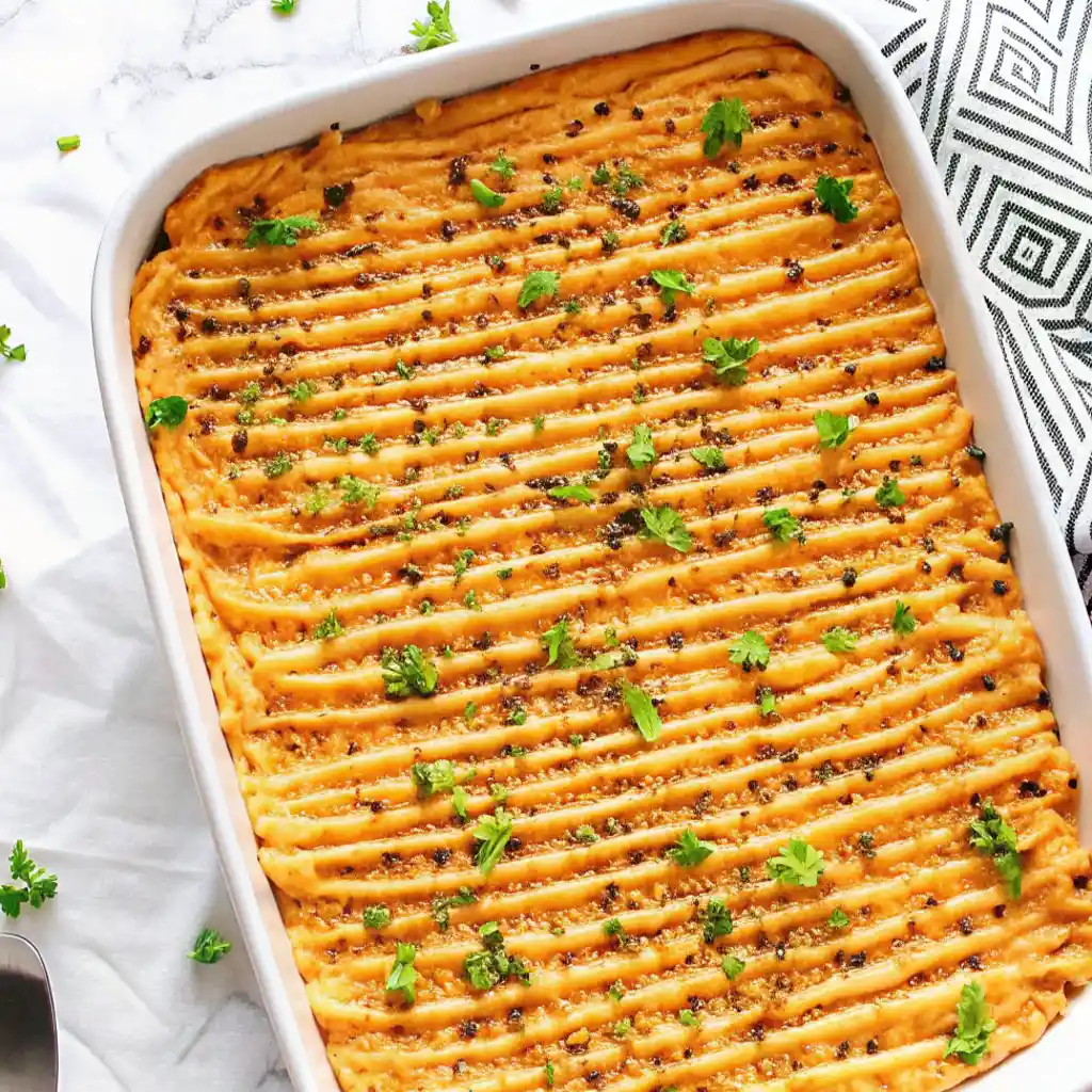 A large white rectangular baking dish holds a salmon sushi bake, featuring a textured, ribbed orange topping garnished with fresh green herbs and dark seasonings, served on a marble surface.