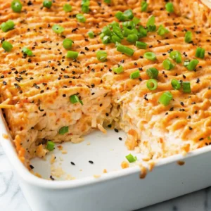 A cross-section of a sushi salmon bake in a white dish reveals layers of rice and creamy salmon, topped with piped spicy mayo, black sesame seeds, and fresh green onions.