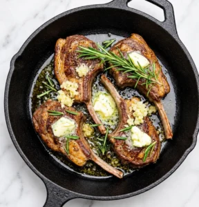 Pan-seared lamb lollipops basting in melted butter with minced garlic and fresh rosemary sprigs inside a black cast-iron skillet, resting on a white marble surface.