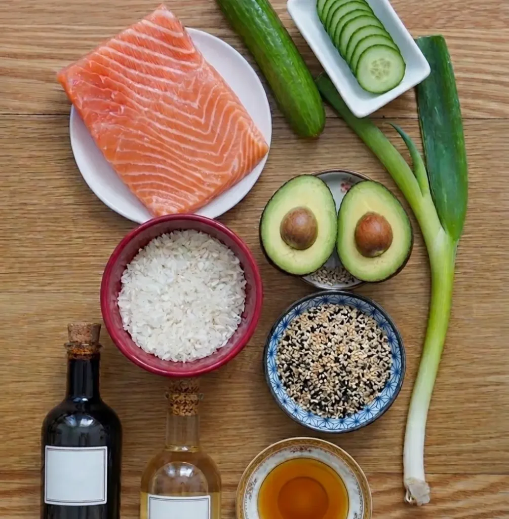 Fresh salmon, white rice, avocado halves, and sliced cucumber arranged on a wooden table with green onions, sesame seeds, soy sauce, rice vinegar, and sesame oil for a poke bowl.