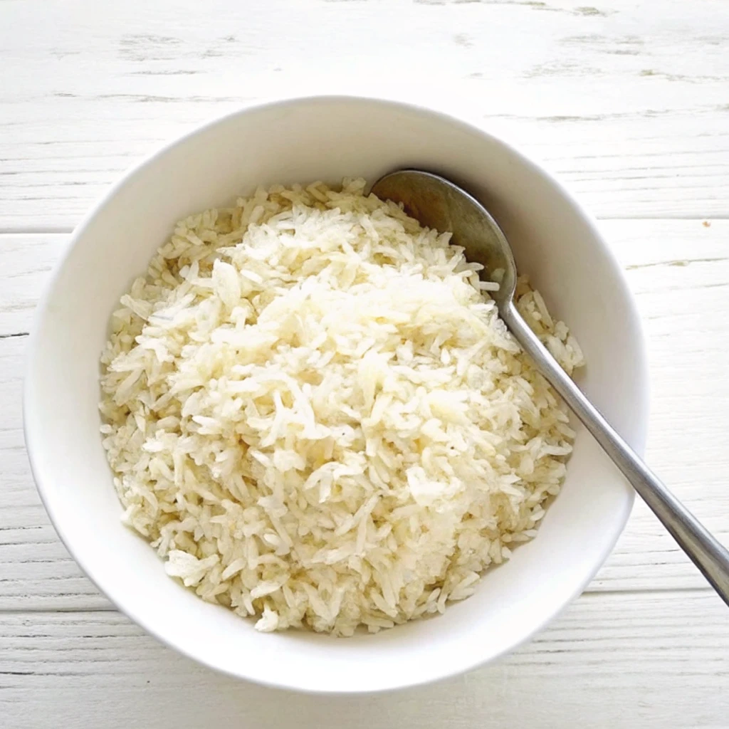 A white bowl filled with fluffy, steamed white rice, featuring a metal spoon resting inside. The bowl is set against a clean, white wooden background for a poke recipe.