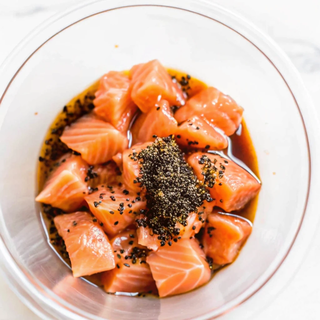Freshly cubed raw salmon sits in a glass bowl, topped with a generous mound of black sesame seeds and a savory liquid marinade, prepared as a base for a poke recipe.