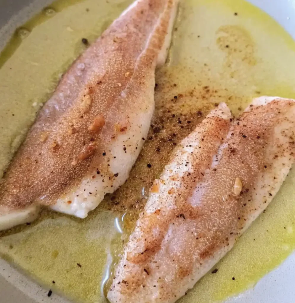 Two golden-brown branzino fillets are sizzling in a shallow pool of olive oil in a light-colored pan. The fish is seasoned with pepper and spices, showing a lovely sear.