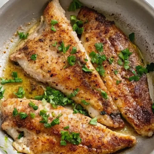Three beautifully searedBranzino Fillet Recipe are arranged in a metal pan, glistening in the butter and oil sauce. They are garnished with a generous sprinkling of fresh, chopped parsley, adding color and aroma to the finished dish.