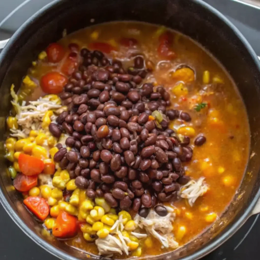 The hearty ingredients for Chicken Taco Soup—including shredded chicken, corn, black beans, and tomatoes—are mixed into a rich, simmering broth in a black pot.