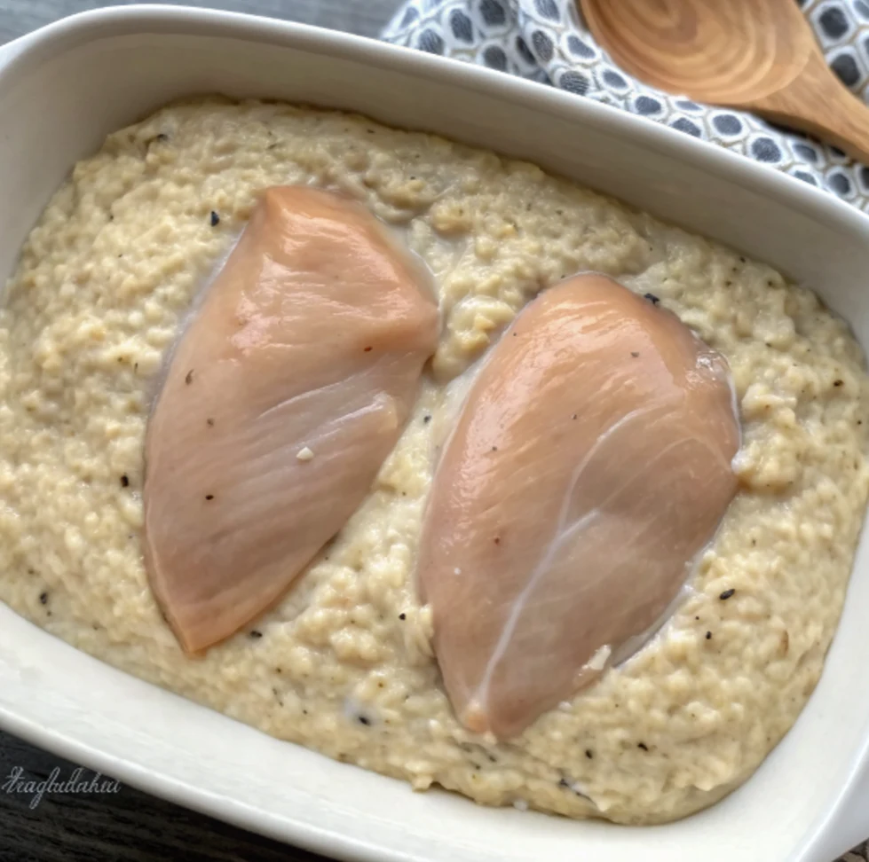 Raw chicken breasts partially submerged in a mixture. The forgotten chicken recipe in a rectangular white baking dish. A wooden spoon and a decorative towel appear in the background.