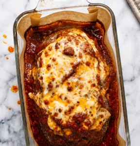 Baked Italian meatloaf in a glass dish, smothered in rich marinara and topped with a thick, bubbling layer of melted golden mozzarella cheese, resting on a marble background.