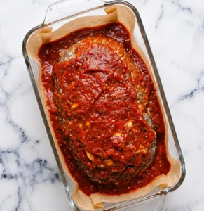 A large Italian meatloaf shaped in a glass baking dish, generously smothered and surrounded by thick, vibrant marinara sauce on a parchment paper lining with a marble counter background.