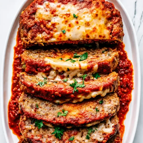 Slices of baked Italian Meatloaf Recipe layered with melted mozzarella cheese and rich marinara sauce on a white platter, garnished with fresh chopped parsley on a marble background.