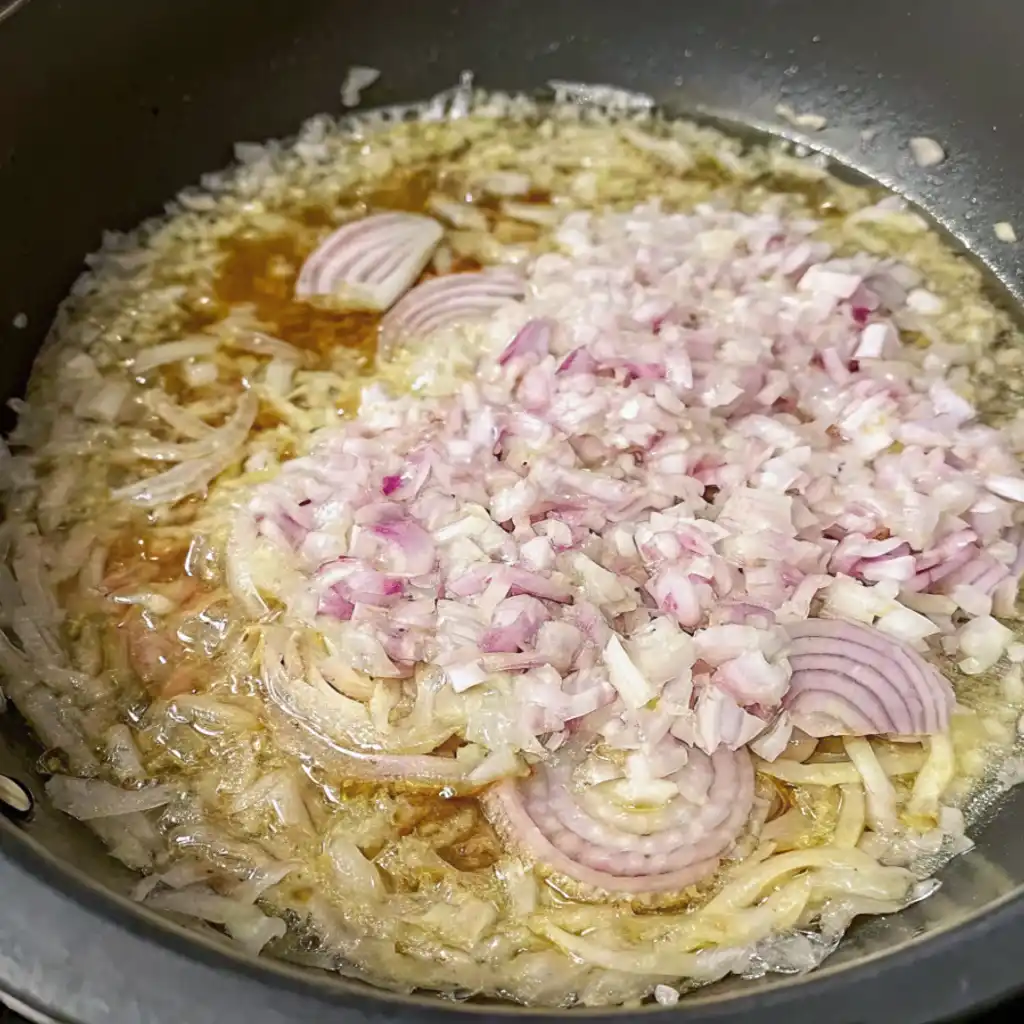 A wide pan is shown from above, with thinly sliced onions and coarsely chopped red onions sauteeing in oil. The onions near the edge are translucent and soft, while the freshly added pieces in the center are still firm and vibrant.