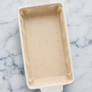 A rectangular white ceramic baking dish lined with beige parchment paper, ready for a recipe, resting flat on a white marble countertop.