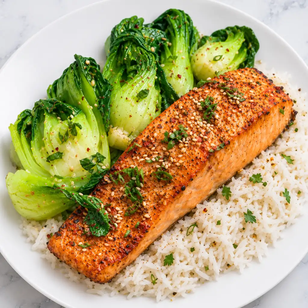 Salmon Asian Rub Recipe Salmon fillet cooked with an Asian spice rub, sprinkled with sesame seeds and herbs, served on a bed of white rice with sautéed bok choy on a white plate.