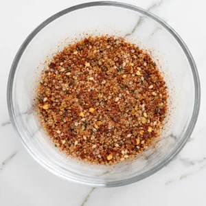 Asian spice blend for salmon, consisting of sesame seeds, chili flakes, and ground spices, presented in a clear glass bowl resting on a marble surface.
