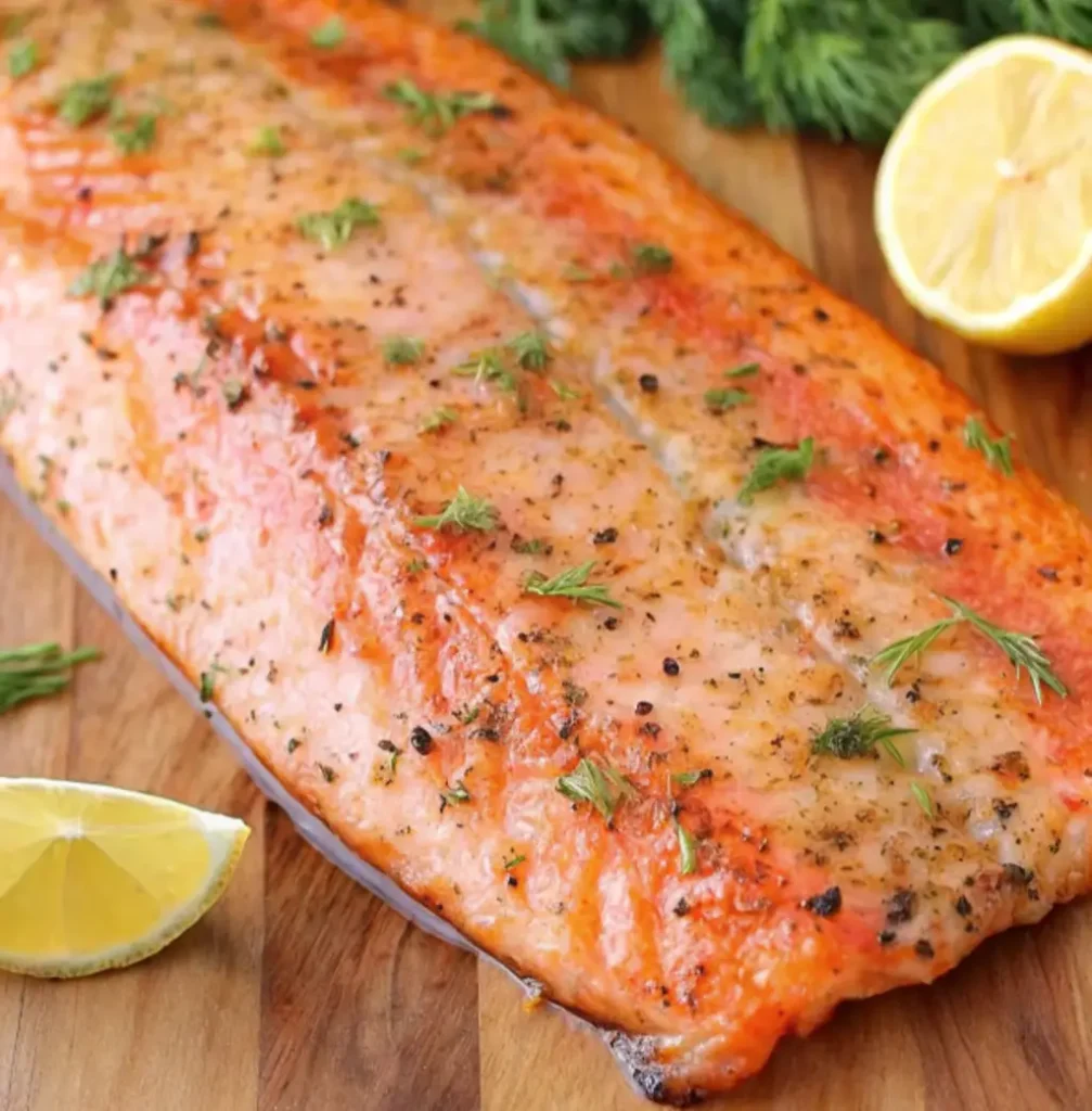A large, golden-brown steelhead trout fillet, cooked to perfection, is presented on a wooden board. It's garnished with fresh dill and flanked by slices of bright yellow lemon, ready to be served.