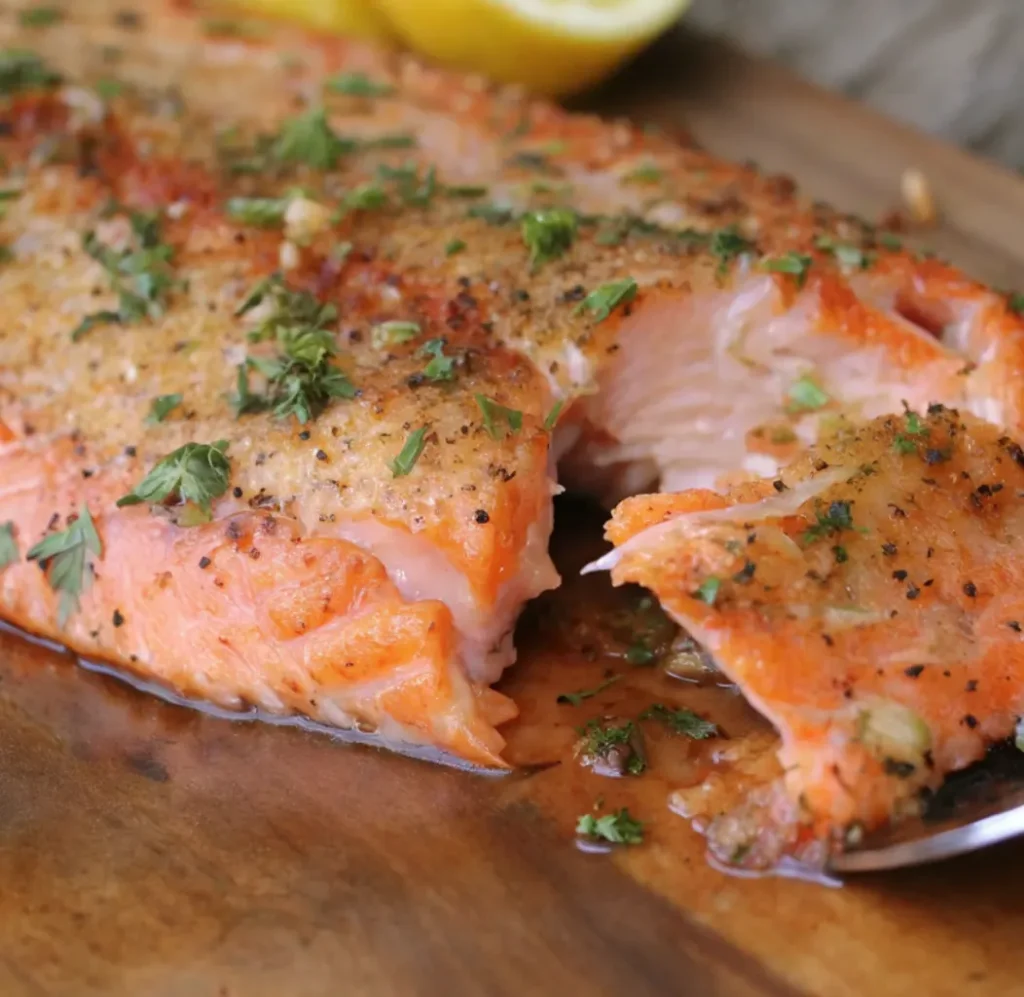 A close-up of a beautifully cooked steelhead trout fillet, flaking easily with a fork, showing its moist, reddish-orange flesh and a golden-brown seasoned crust of herbs and spices, resting on a wooden board.