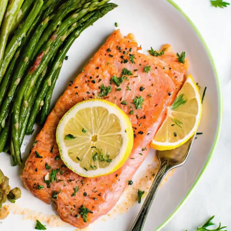 A plated meal of perfectly cooked steelhead trout Recipe fillet, garnished with fresh lemon slices and parsley, served alongside a vibrant green bundle of roasted asparagus, ready to be eaten.