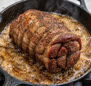 Tied seasoned venison roast seared to a golden-brown crust in hot, sizzling oil inside a cast-iron skillet.