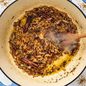 A wooden spoon stirring caramelized onions, whole cinnamon sticks, and mustard seeds cooking in oil inside a Dutch oven for a lamb vindaloo base