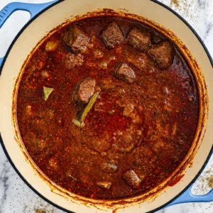 A top-down view of fully cooked lamb vindaloo with tender meat submerged in a rich, dark red spicy curry sauce in a Dutch oven.