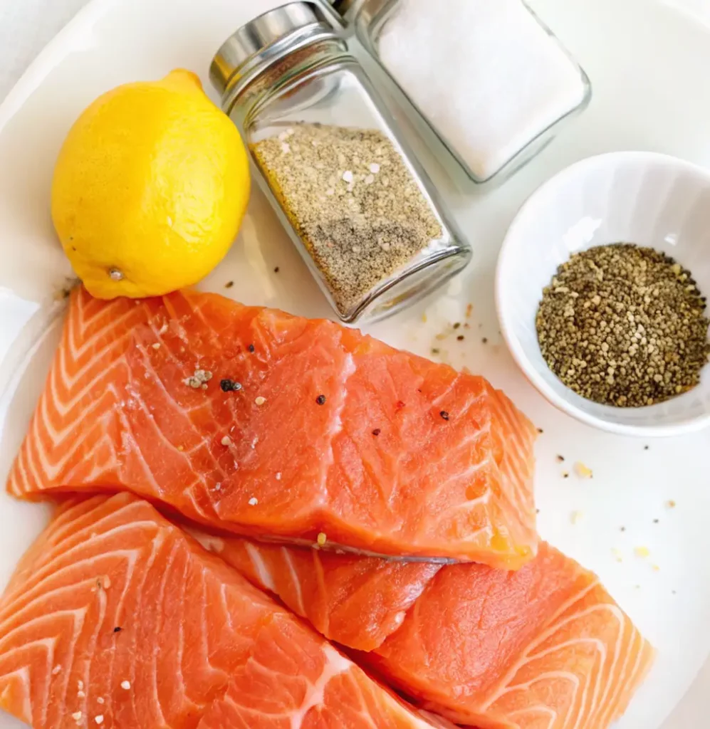 3 vibrant orange salmon fillets on a white plate with a whole lemon, shakers of salt and pepper, and a small white bowl of ground black pepper nearby.