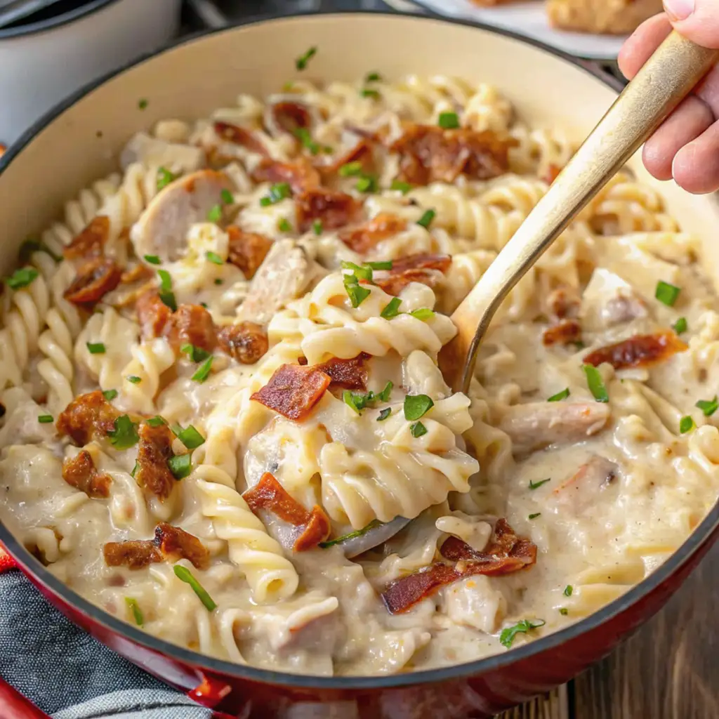 A creamy Chicken Bacon Ranch Pasta with tender chicken pieces, crispy bacon bits, and a rich ranch-flavored sauce, garnished with fresh chives, being scooped from a red skillet.