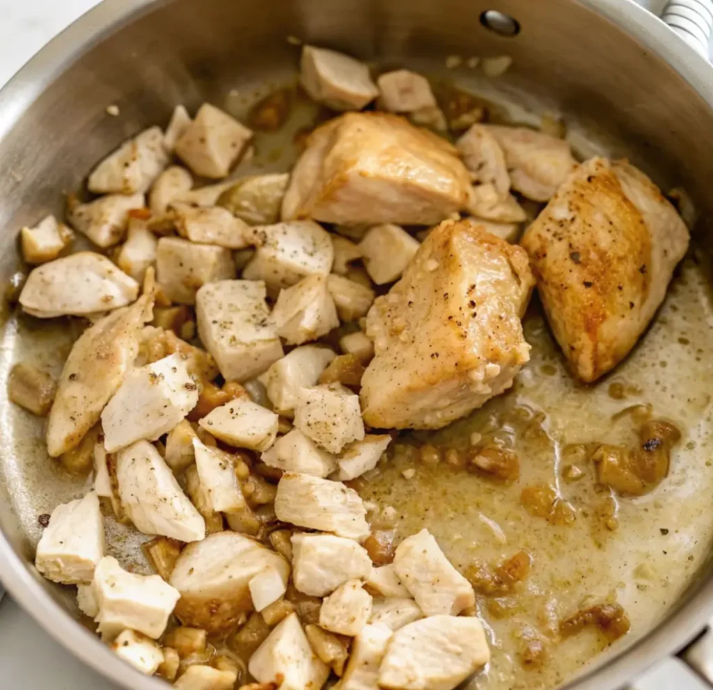 Cooked chicken breast pieces, some diced and some larger, are sautéing in a stainless steel skillet with rendered fat and seasonings,