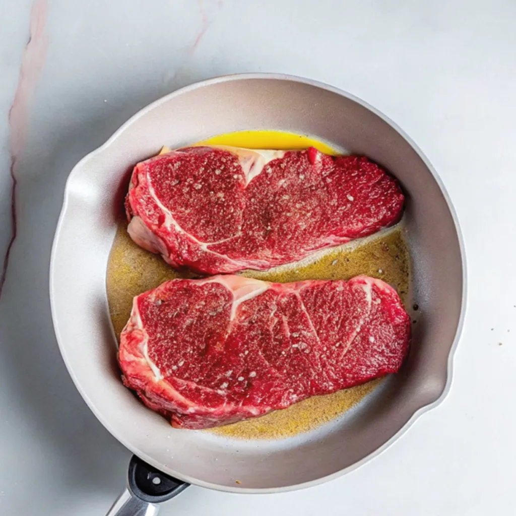 Two raw, seasoned chuck steaks are placed in a light gray frying pan with hot oil, ready to be seared