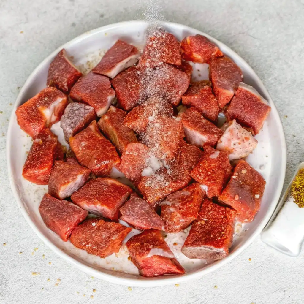 Seasoning Raw Steak Cubes: A white plate holds raw beef cubes, being seasoned with salt and pepper from above, preparing them for cooking in the garlic butter sauce.
