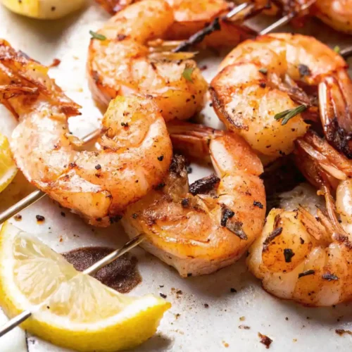 A close-up of several smoked shrimp skewers, showing the pink-orange color and seasoning, next to a lemon wedge on a light-colored surface, highlighting a finished dish.