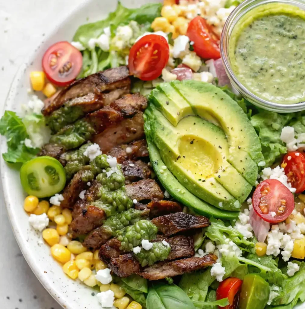 A vibrant steak salad in a white bowl, featuring sliced grilled steak drizzled with green dressing, mixed greens, corn kernels, cherry tomatoes, crumbled cheese, and a halved avocado.