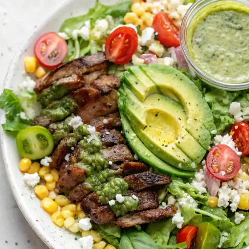A vibrant steak salad in a white bowl, featuring sliced grilled steak drizzled with green dressing, mixed greens, corn kernels, cherry tomatoes, crumbled cheese, and a halved avocado.