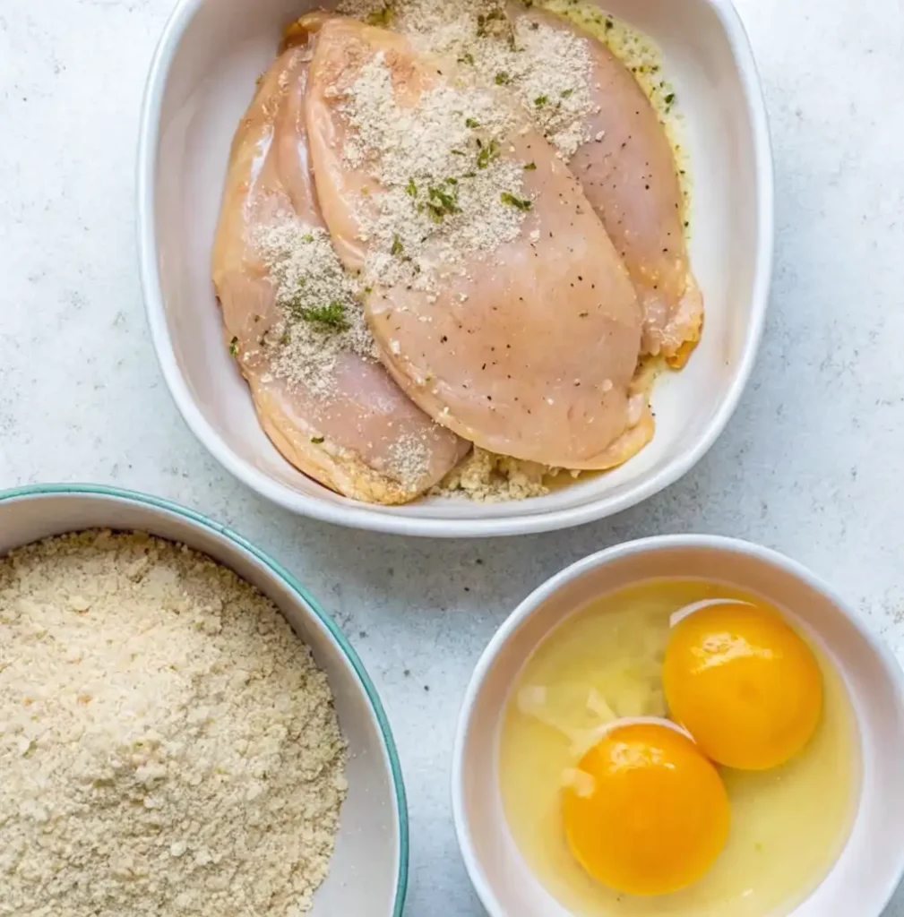 Raw chicken breasts (or cutlets) are shown, coated in a breading mixture, next to a bowl of breadcrumbs and another with two cracked eggs, ready for an easy baked chicken cutlet recipes.