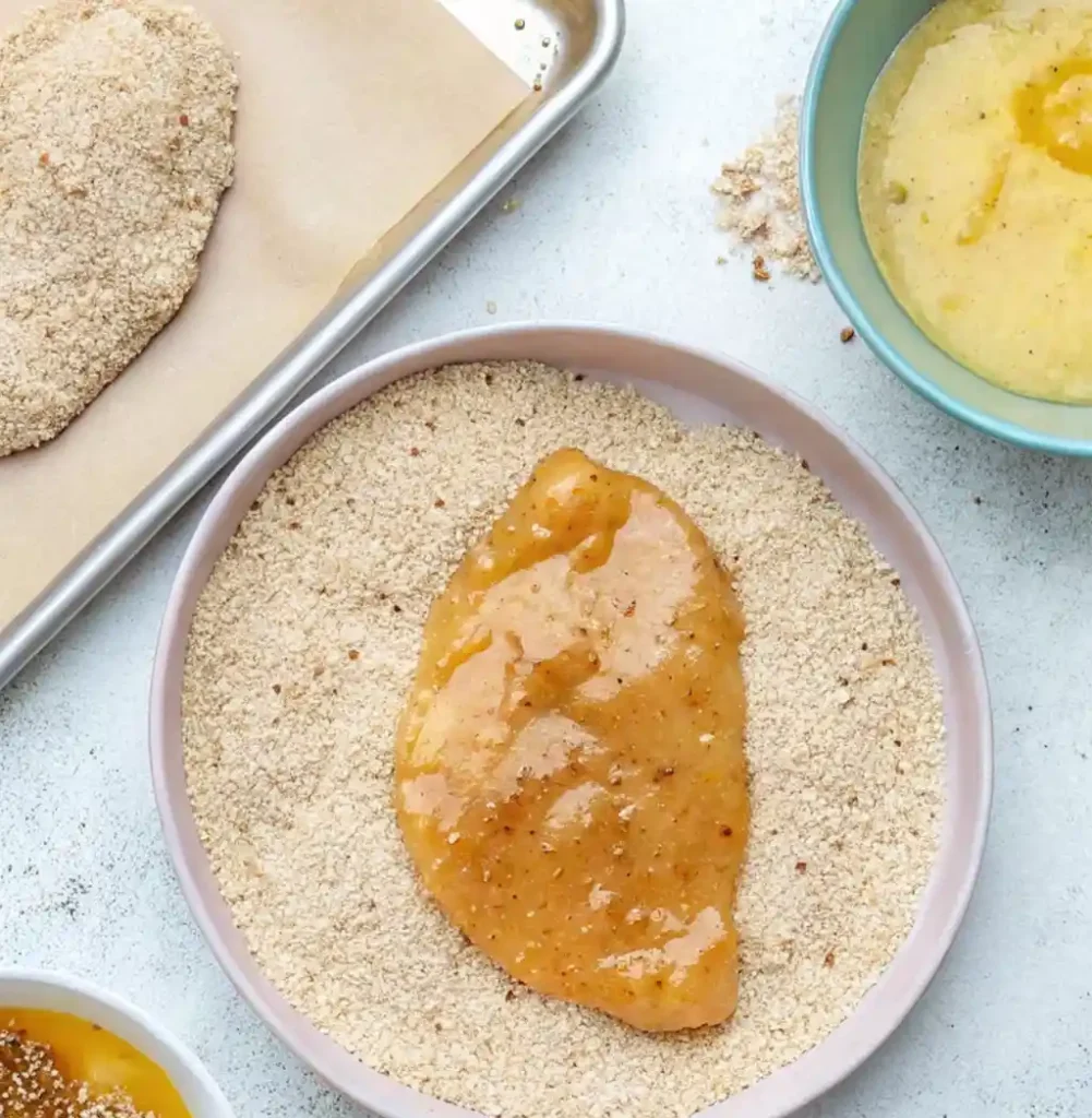 A chicken cutlet, already dipped in an egg and spice mixture, is being pressed into a shallow dish of breadcrumbs to finish its coating before being baked on a parchment-lined tray.