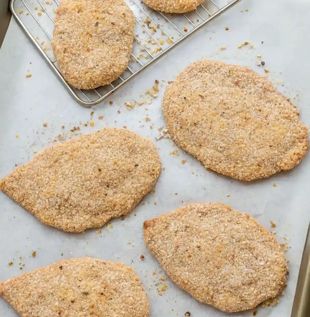 Several breaded chicken cutlets are laid out on a parchment-lined baking sheet, ready for the oven. One is resting on a wire rack, which helps to circulate heat for an extra crispy baked coating.