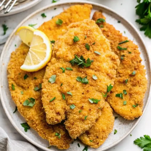 three crispy, golden-brown baked chicken cutlets stacked on a white plate, garnished with fresh parsley and a bright wedge of lemon. The texture shows a light, crunchy breading. for baked chicken cutlet recipes