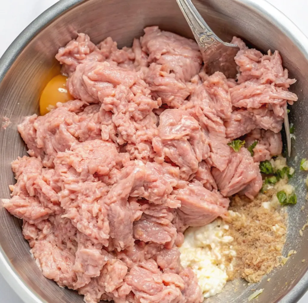 A metal mixing bowl containing the raw ingredients for chicken meatloaf recipe, including coarse ground chicken, a cracked egg yolk, breadcrumbs, ricotta cheese, and chopped green herbs.