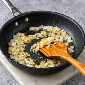Chopped onions and minced garlic sauté in oil inside a black skillet, stirred with a wooden spatula
