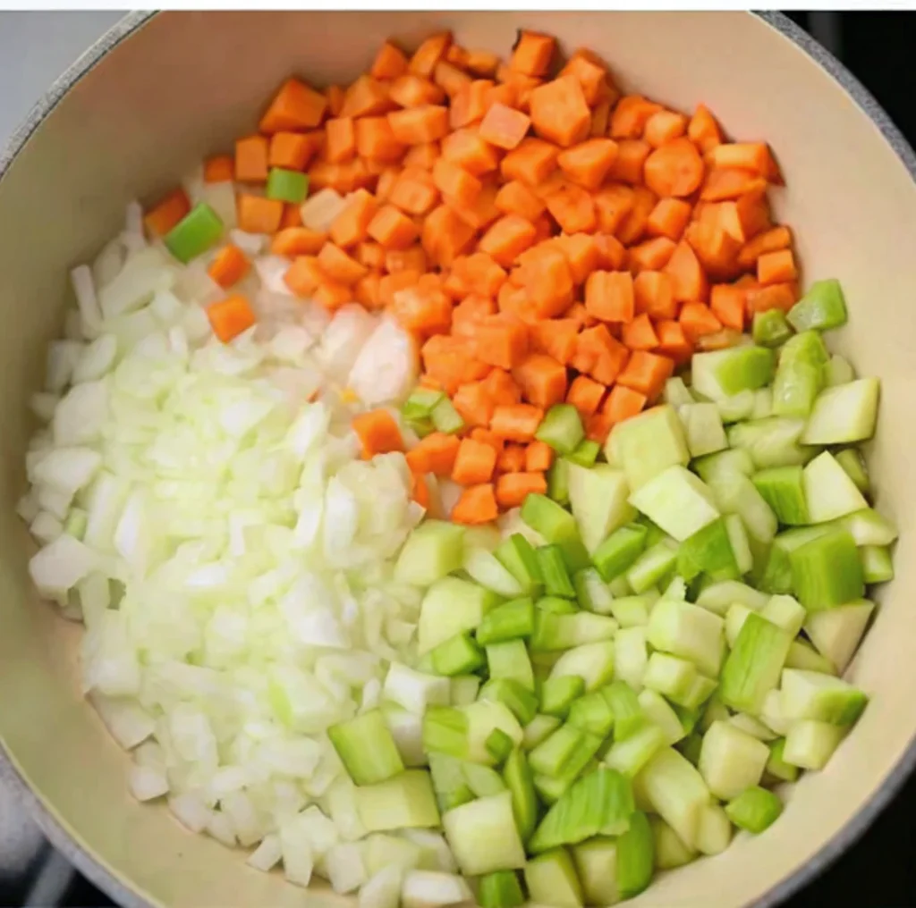 diced vegetables carrots, white onions, and celery separated into three piles in a light-colored pot, ready for sautéing to form the base for a soup.