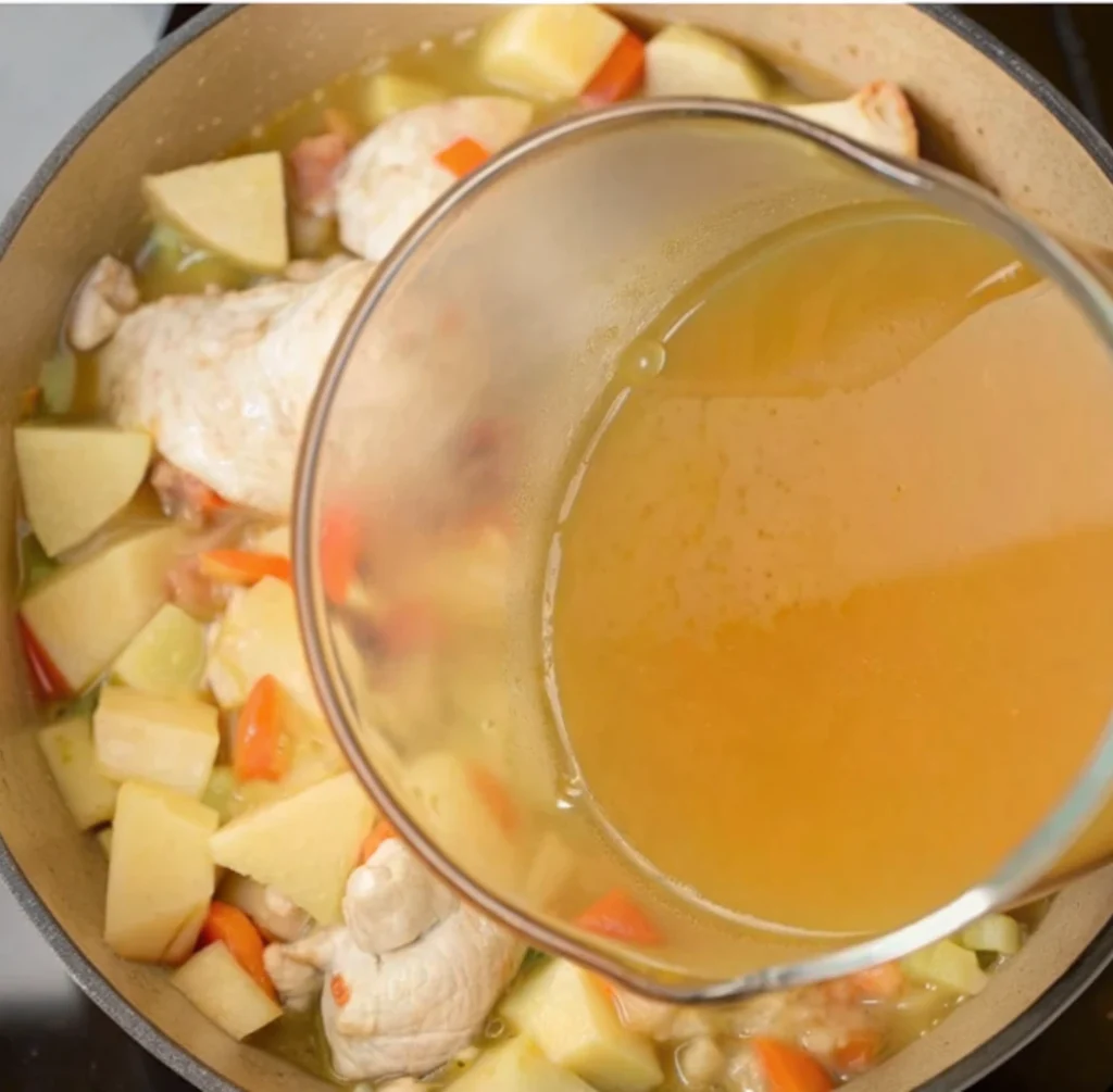 A glass measuring cup is used to pour light-brown broth into a pot containing partially cooked chicken pieces, diced carrots, celery, and large chunks of potato, combining the soup ingredients.