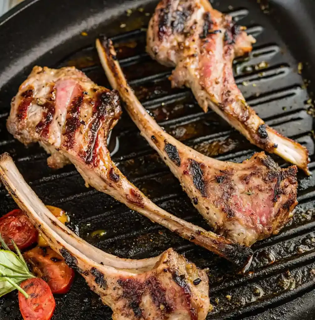 Four well-seared Grilled Lamb Chops are shown in a black cast iron grill pan. The chops feature distinct dark grill marks and are served alongside a few charred tomato slices and a sprig of basil or rosemary.