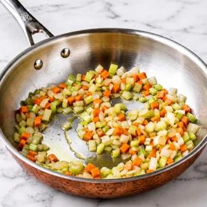 Finely diced carrots, celery, and onions sautéing in oil inside a stainless steel skillet with a copper base, resting on a white marble countertop.