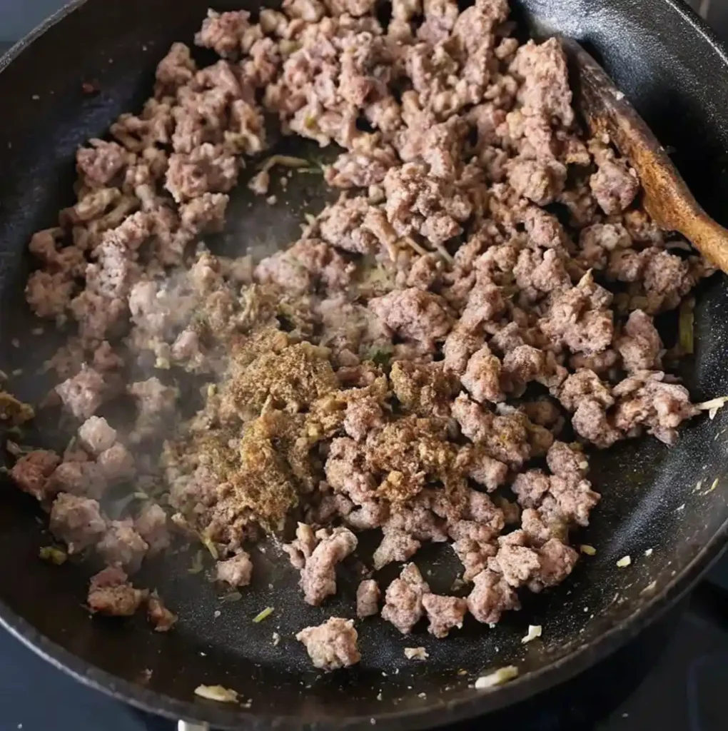 Ground lamb is shown browning and cooking in a black skillet, with a scoop of spices being added to the meat, which is being stirred with a wooden spoon for the Bolognese sauce.